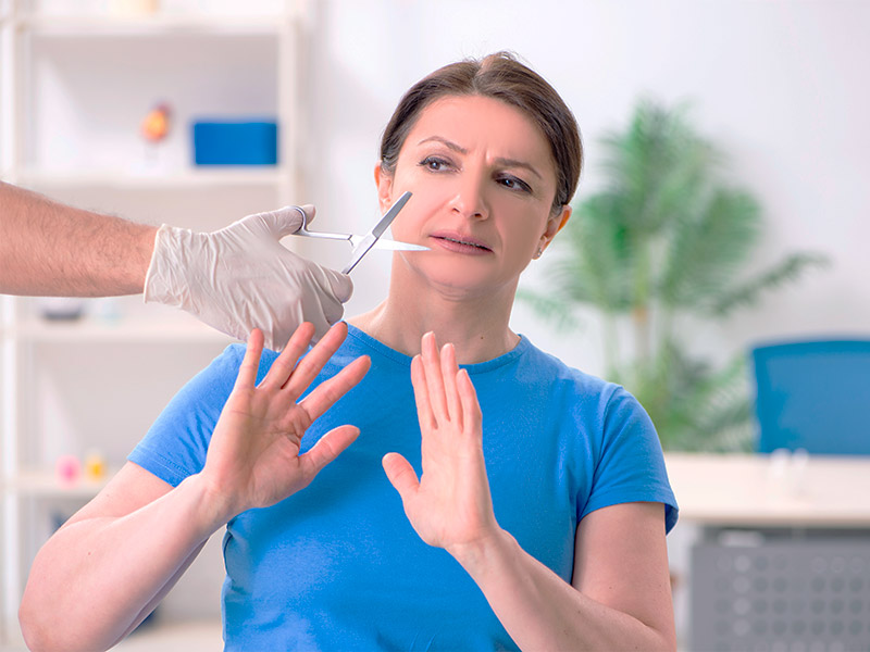 Mão com luva com uma tesoura na frente do rosto de mulher, que foge de uma cirurgia plástica que deu errado