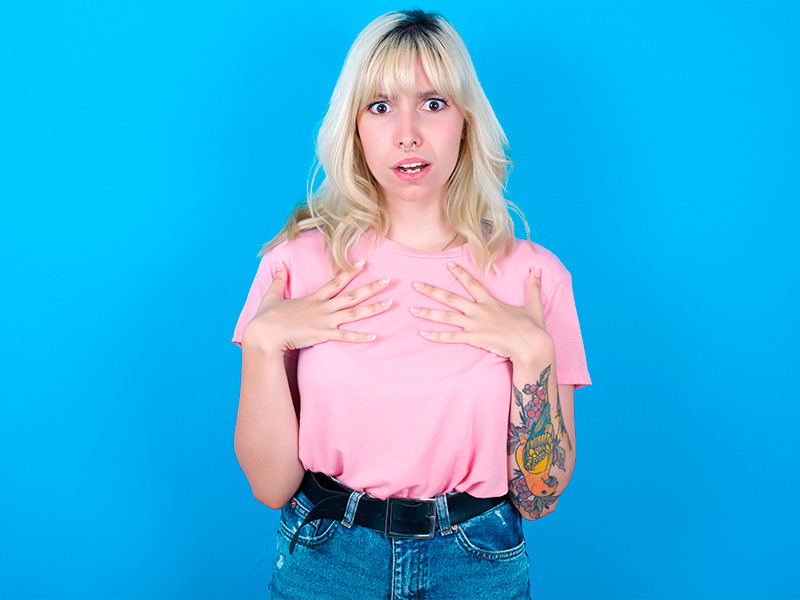 Mulher de camiseta rosa e calça jeans, com a mão no peito, com a boca aberta assustada que pode ter queloide na tatuagem em seu braço esquerdo