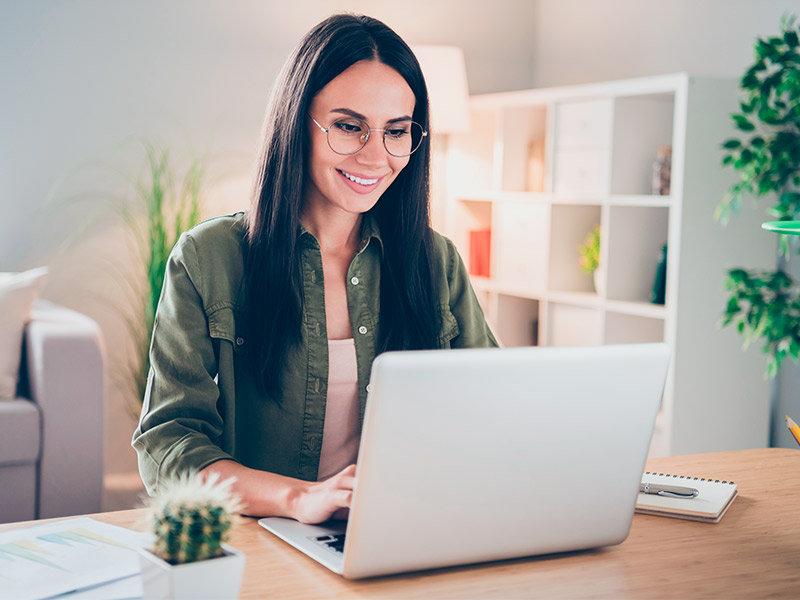 Como evitar queloide? Mulher de camisa verde e óculos, sentada na mesa da sala sorrindo enquanto pesquisa como evitar queloide no notebook