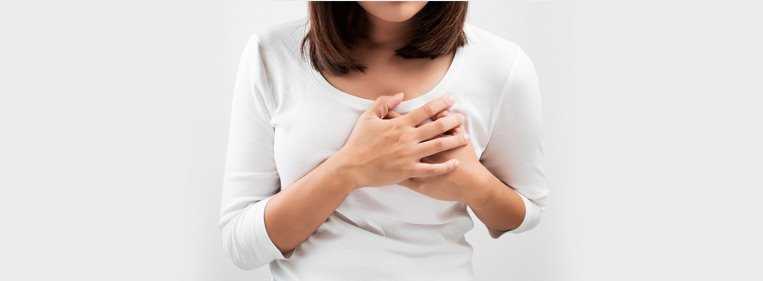 Mulher de camiseta branca de manga longa com a mão no peito com falta de ar, um dos sintomas da embolia pulmonar