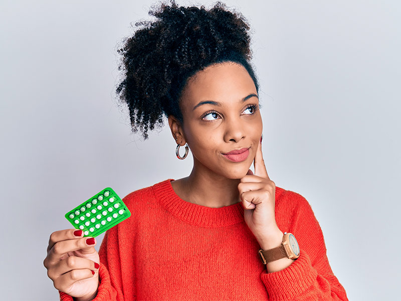 Mulher olhando para o lado com a mão no queixo, segurando uma cartela de pílulas, pensando se o anticoncepcional causa trombose