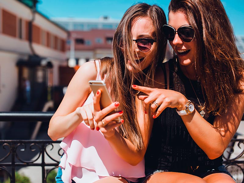 Na imagem, duas mulheres estão sorrindo e olhando para a tela do celular enquanto procuram por fotos de resultados de rinoplastia