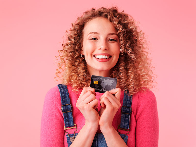 Imagem com fundo rosa e uma mulher em primeiro plano, segurando um cartão de crédito