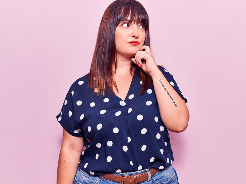 A cirurgia de gigantomastia é a solução para diminuir seios enormes? Mulher de camisa azul com bolinhas brancas e seios enormes, com a mão esquerda no rosto pensando se faz uma cirurgia de gigantomastia
