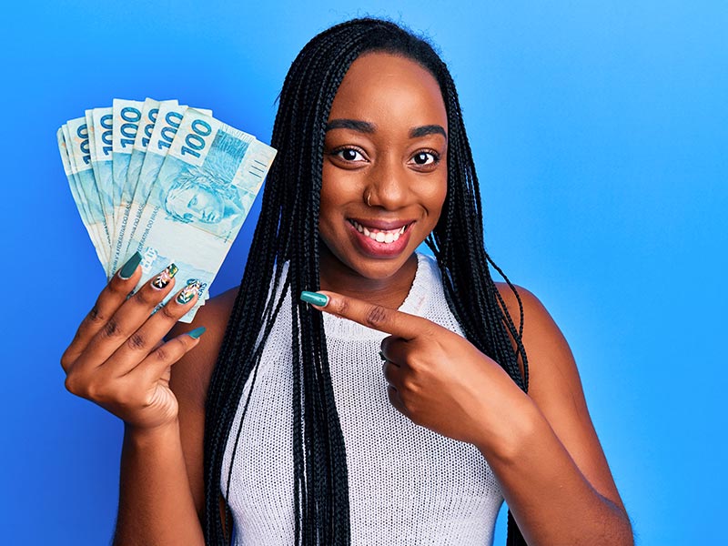 Mulher sorrindo, segurando várias notas de 100 reais em uma mão e apontando com a outra para pagar o preço da gigantomastia 