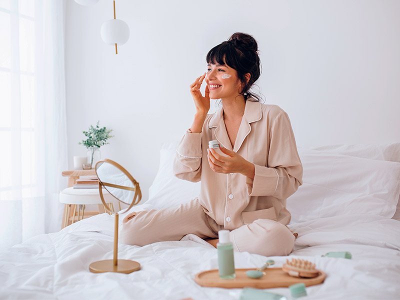 Imagem de uma mulher sentada na cama, passando creme facial no rosto, uma das possibilidades para suavizar rugas nos olhos 