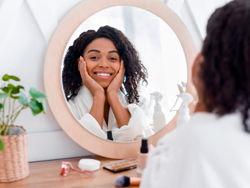 Imagem de uma mulher se olhando no espelho com expressão de felicidade após saber os benefícios do ácido hialurônico para o rosto