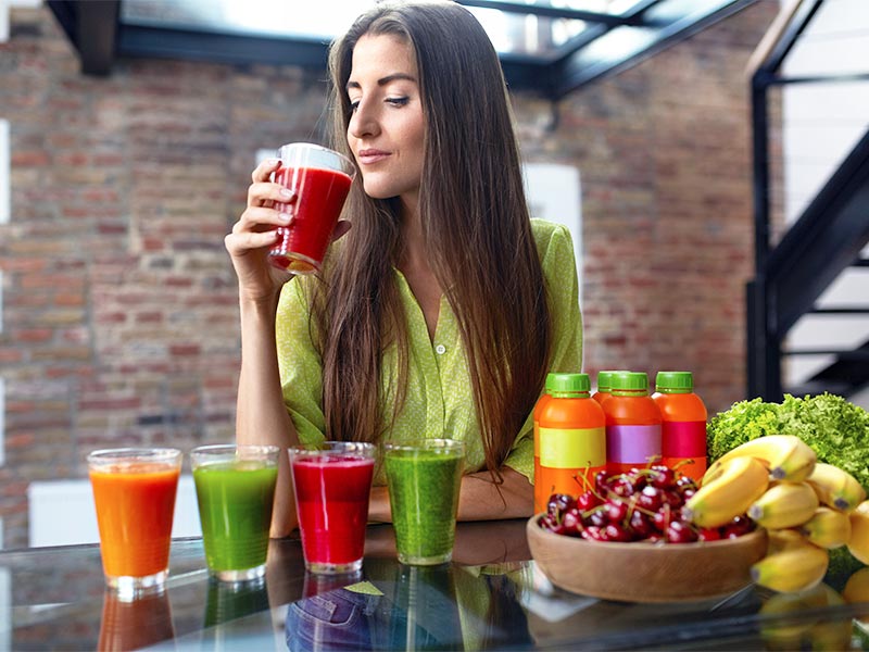 Mulher tomando suco para rejuvenescer, com outros 4 copos de suco, três garrafas e uma cesta de frutas na sua frente