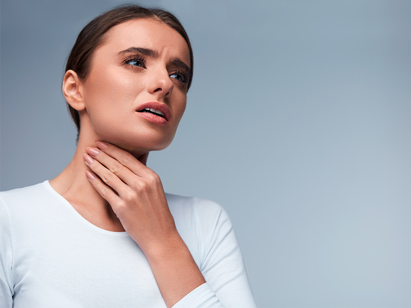 Caroço dolorido pode ser lipoma? Mulher de camiseta branca de mangas compridas, com sintomas de lipomas, com a mão no pescoço
