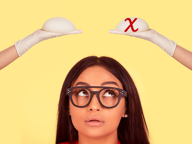 Mulher de óculos olhando para cima, onde tem duas mãos com luva segurando uma prótese de silicone, uma delas com um X em cima para mostrar que ela está na dúvida entre a mastopexia com ou sem prótese