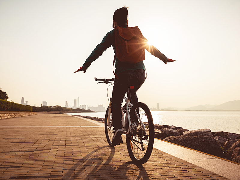 Exercícios para diminuir os seios funcionam? Mulher andando de bicicleta na orla da praia, fazendo exercícios para perder peito
