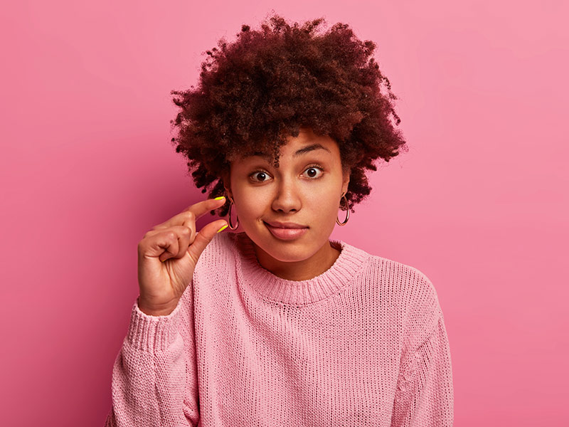 mulher em um fundo rosa, ela faz o sinal de "pequeno" com as mãos., demonstrando as poucas chances de ter contratura