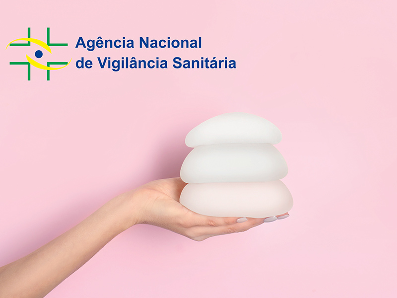 imagem com fundo rosa, uma mão segurando 3 próteses de silicone. O logo da ANVISA está ao canto.