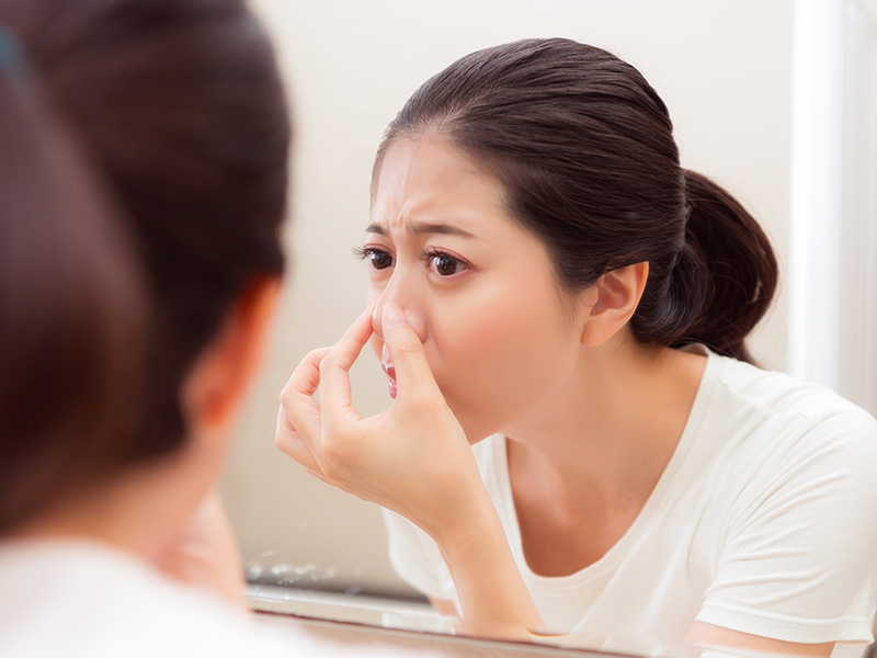 Uma jovem se questionando com quantos anos pode fazer rinoplastia, observa a própria imagem no espelho enquanto toca a ponta do nariz