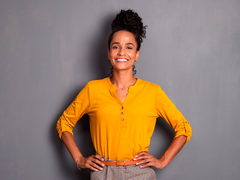 Imagem com funda cinza e uma mulher negra, de cabelos cacheados e preso, vestindo uma calça xadrez e uma blusa amarela, sorrindo em primeiro plano