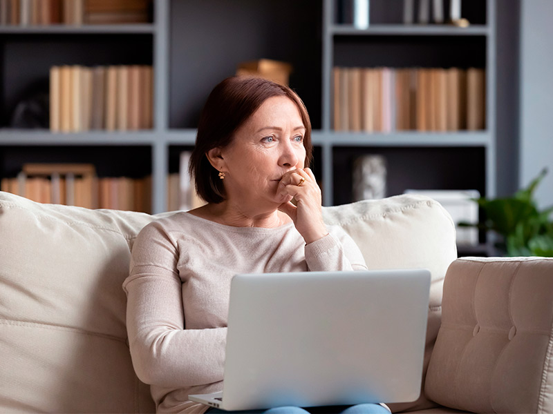 Mulher de meia idade sentada no sofá, com um notebook no colo pesquisando sobre a recuperação da blefaroplastia