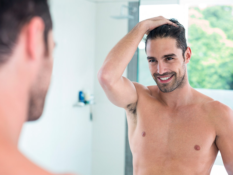 Homem passando uma mão no cabelo sorrindo enquanto se olha no espelho depois da cirurgia de mama masculina