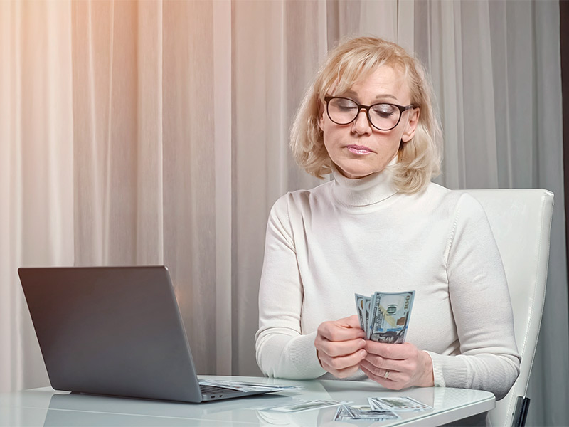 Imagem de uma mulher segurando notas de dinheiro e pesquisando no notebook o preço do botox 