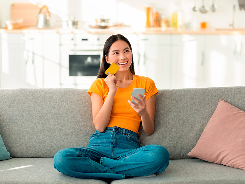 Foto de uma mulher sentada em um sofá cinza, com expressão de felicidade ao pesquisar o valor da cirurgia de testa no celular. A modelo também está segurando um cartão de crédito. 