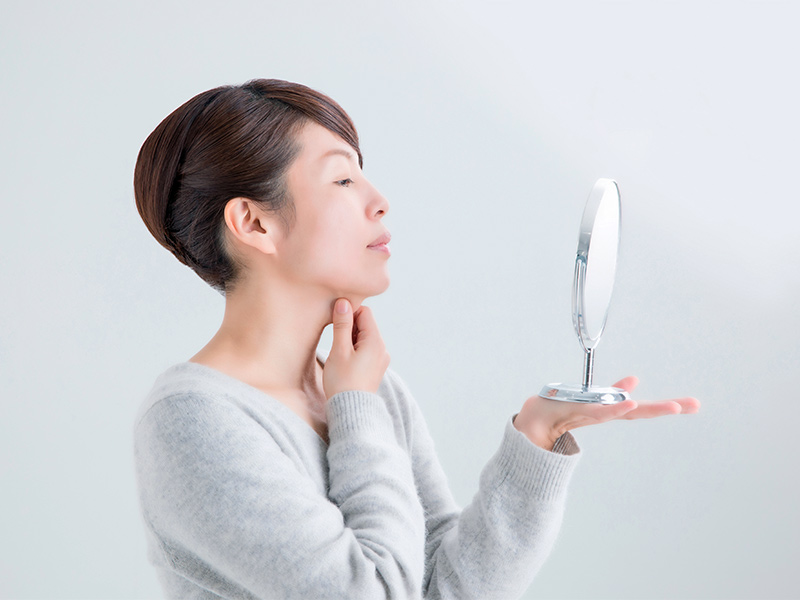 Imagem de uma mulher pensando em realizar um lifting de pescoço, enquanto se olha no espelho e segura a gordura localizada na região da papada 