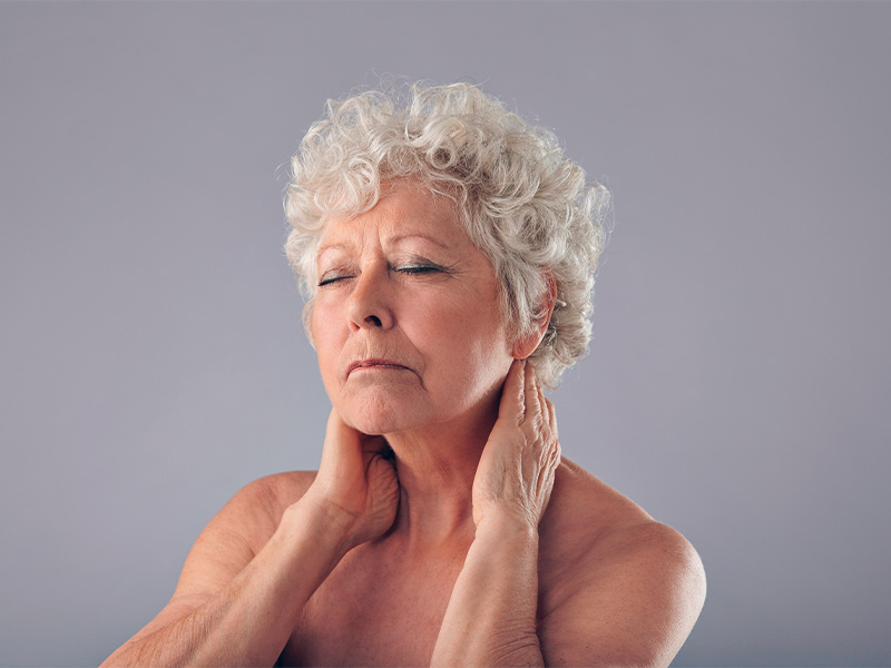 Imagem de uma mulher na terceira idade, com as mãos ao redor do pescoço e expressão de cansaço enquanto pensa sobre o lifting de malar para melhorar o contorno facial