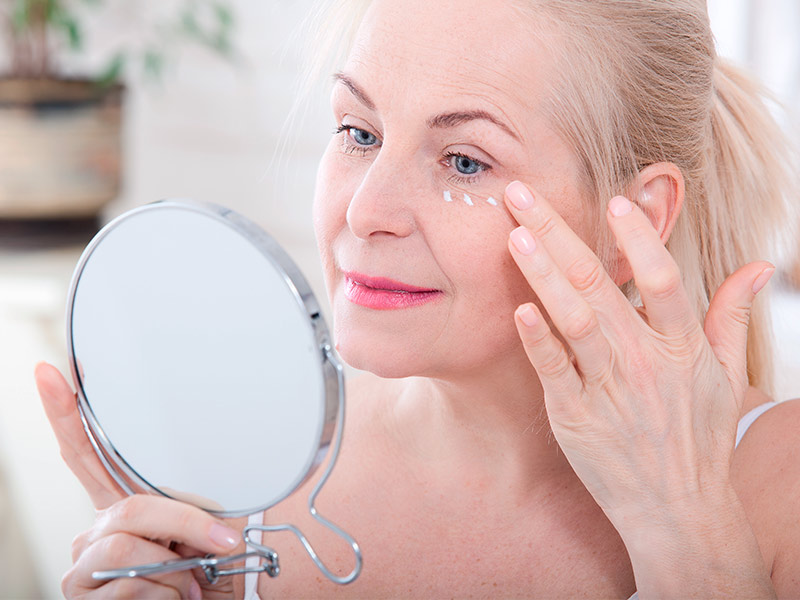 Imagem de uma mulher se olhando no espelho e passando creme na região dos olhos para conseguir o efeito lifting 