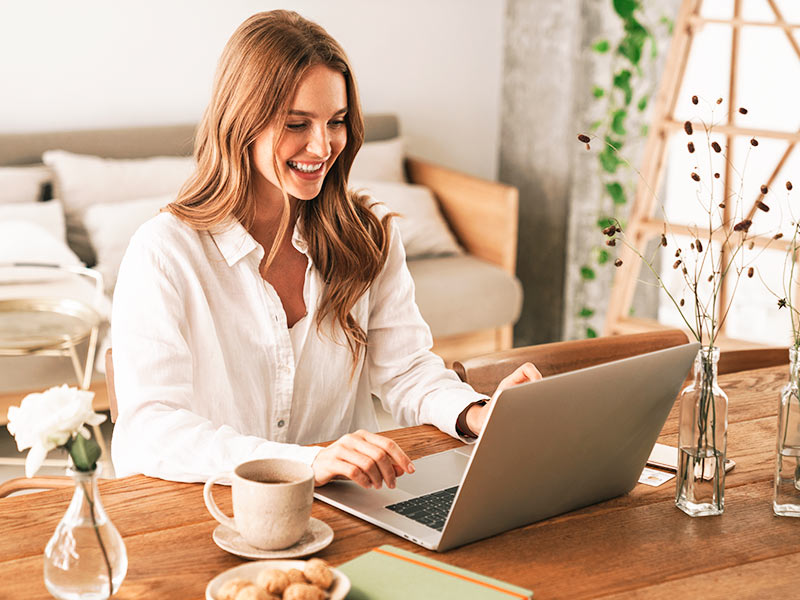 Mulher sentada na mesa sorrindo enquanto pesquina no notebook a cirurgia para tirar a pele e gordura dos flancos