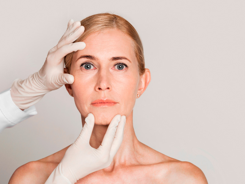 Imagem do rosto de uma mulher de meia idade com rugas e as mãos de um especialista fazendo uma avaliação em sua pele 