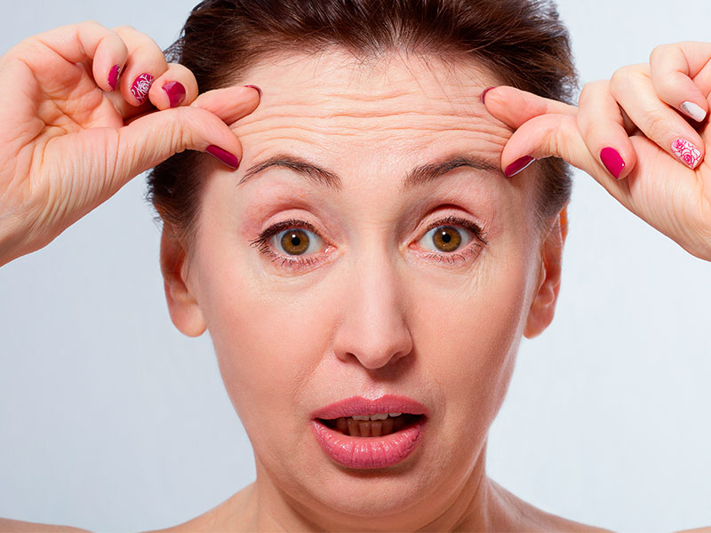 Imagem de uma mulher de meia idade, com olhos arregalados e boca aberta, enquanto coloca as mãos na testa para destacar as rugas