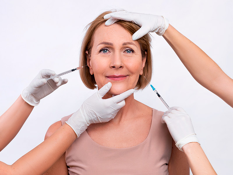 Imagem de uma mulher de meia idade sorrindo, enquanto duas profissionais seguram seringas de preenchimento próximo ao seu rosto