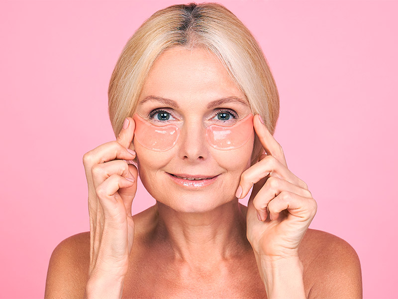 Imagem com fundo rosa e uma mulher de meia idade em primeiro plano. A modelo está sorrindo e usa dois adesivos anti rugas, um de cada lado do rosto, abaixo dos olhos. 