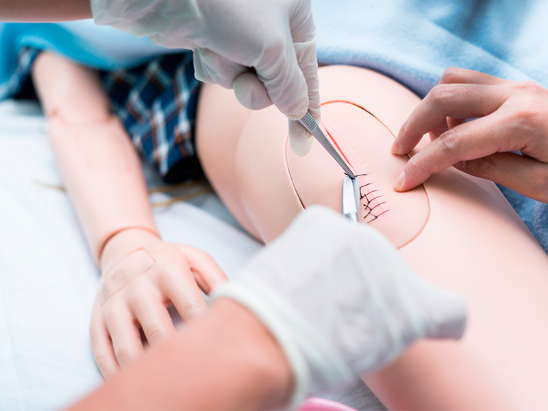 Mãos com luva mostrando como tirar pontos de cirurgia plástica no joelho de boneco