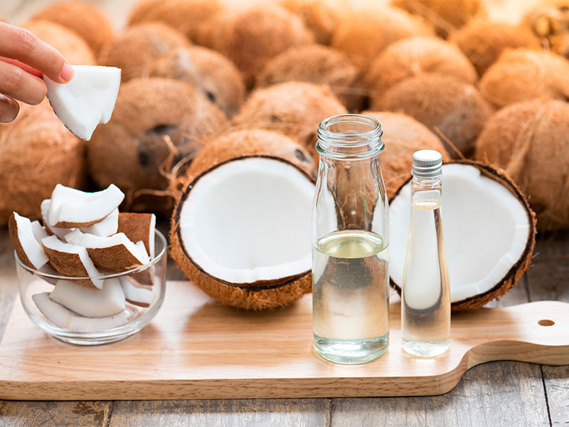 Imagem de uma mesa em primeiro plano, com uma tábua de corte e pedaços de coco em cima, além de dois recipientes de óleo de coco para marcas de expressão. 