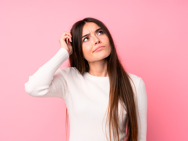 Pode fazer mastopexia com quantos anos? Mulher morena, de camiseta de manga longa branca, com a mão direita na cabeça, olhando para cima e pensando com quantos anos pode fazer a mastopexia