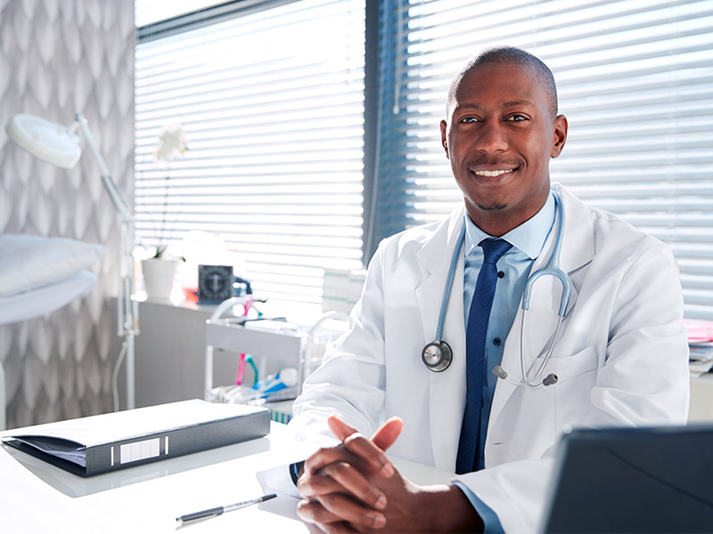 Na foto um médico aparece sorridente, em seu consultório. Os dermatologistas e cirurgiões plásticos são quem pode fazer harmonização facial.