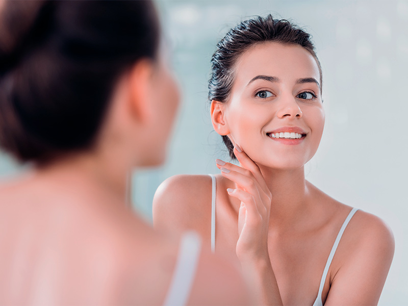 Mulher se olhando no espelho sorrindo, com a mão na mandíbula pensando em quanto tempo dura o preenchimento do bigode chinês