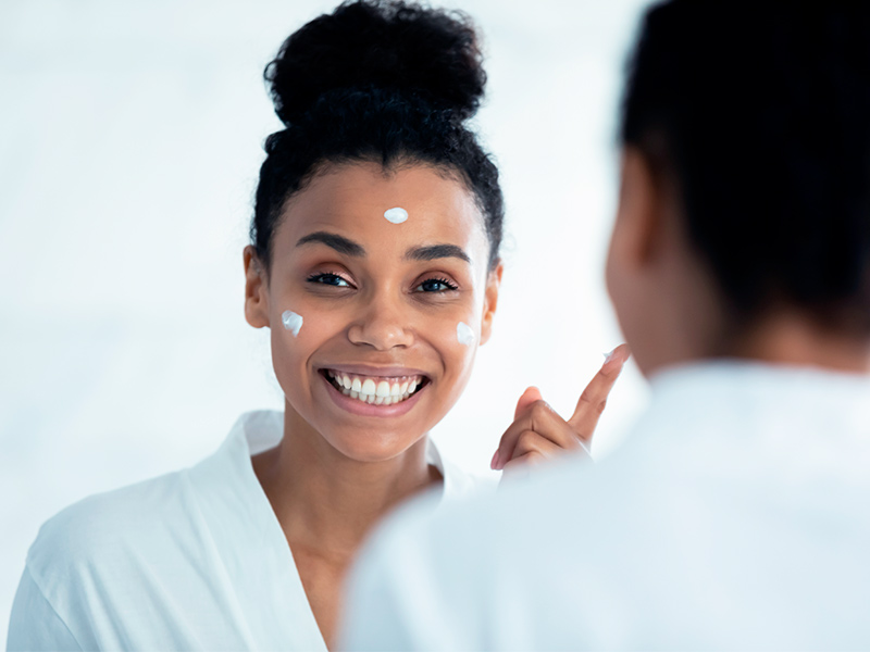 Foto de uma mulher se olhando no espelho e passando hidratante facial, um dos principais cuidados após harmonização facial