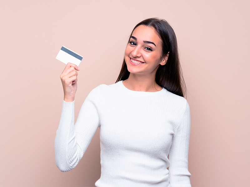 Imagem com fundo rosa claro e uma mulher em primeiro plano, segurando um cartão de crédito