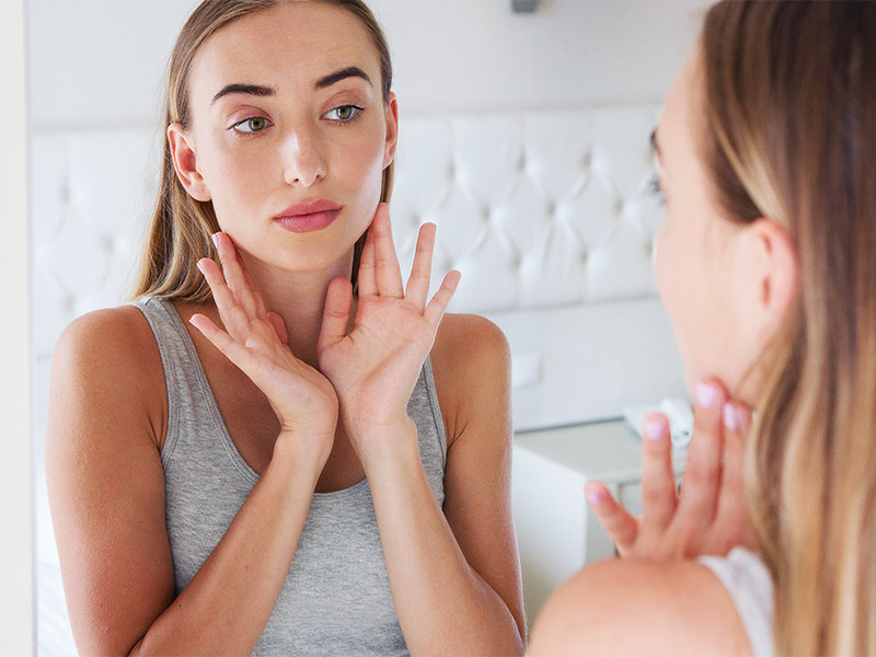 Imagem de uma mulher se olhando no espelho e analisando se o rosto está inchado após aplicação do bioestimulador de colágeno