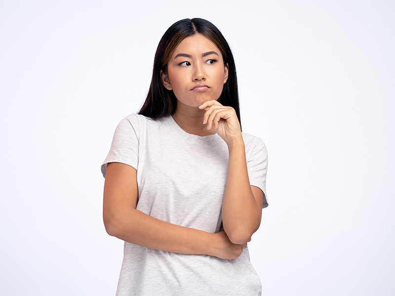 Mulher de camiseta branca, com a mão esquerda no queixo, olhando para o lado fazendo biquinho pensando se cisto sebáceo na cabeça é câncer