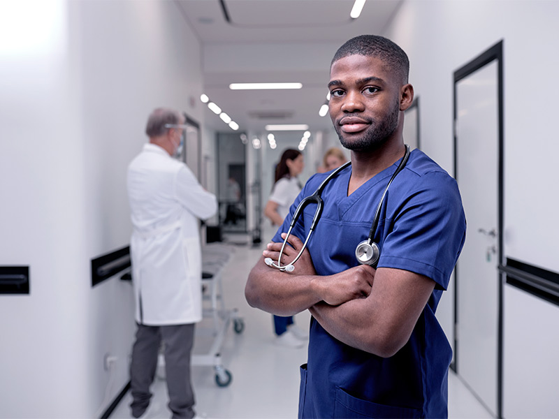 Médico parado no corredor de um hospital com uniforme azul e estetoscópio no pescoço, de braços cruzados