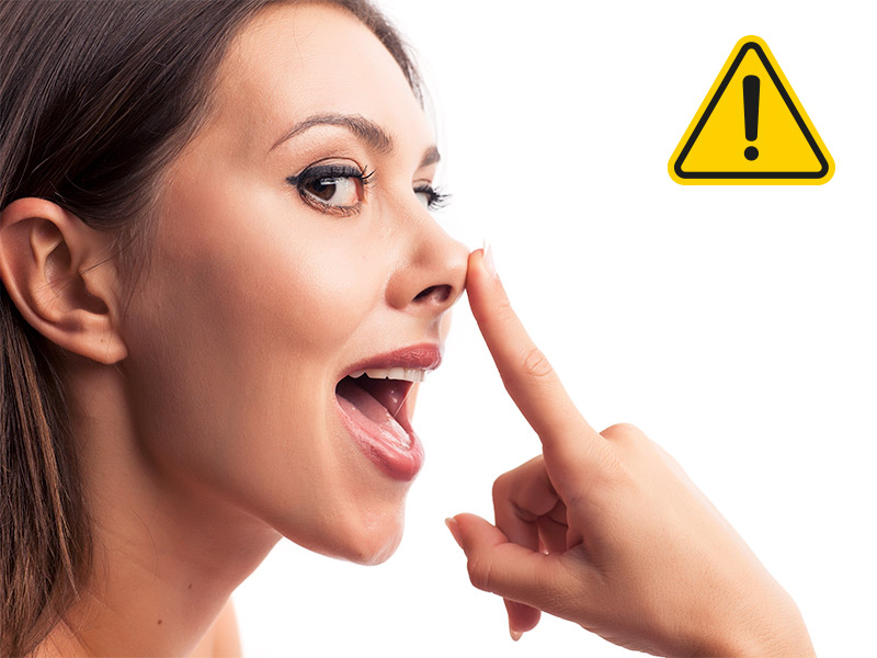 Imagem de uma mulher com a boca aberta e o dedo indicador na ponta do nariz. Do lado direito da foto há um sinal de alerta devido aos riscos do metacril no nariz.