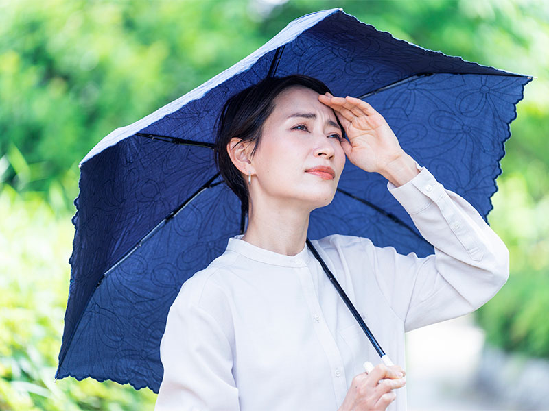 Mulher de camisa branca com a mão na testa, segurando um guarda-chuva azul, para proteger a cicatrização de pequena cirurgia do sol