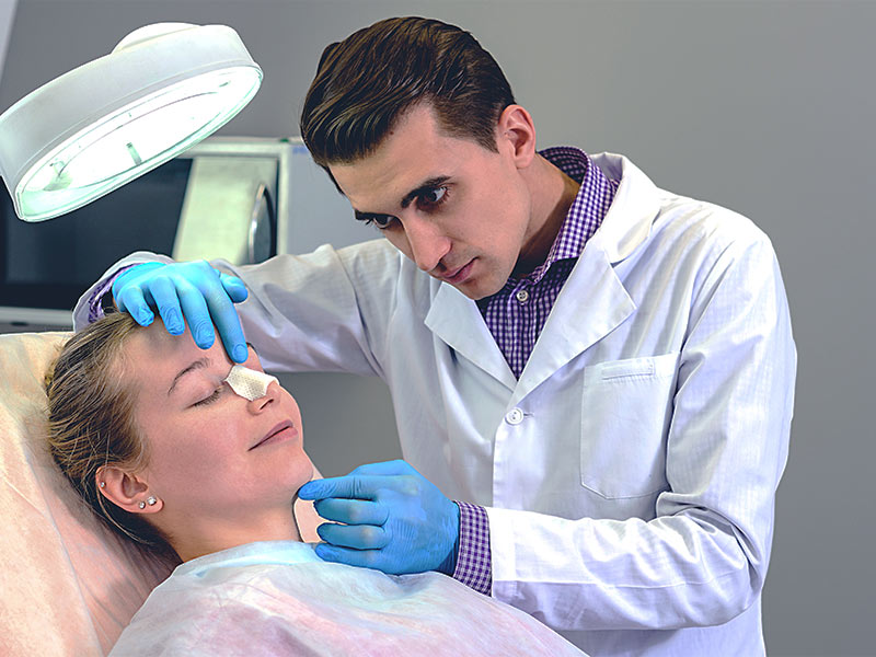 A imagem mostra um médico avaliando o curativo de uma paciente após a realização da cirurgia de rinoplastia