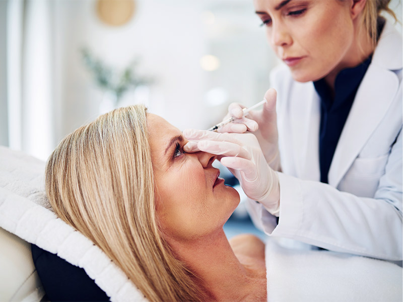 Imagem exemplificando como é feita a aplicação de botox dentro do consultório médico. Na foto, a paciente está deitada enquanto a médica aplica a substância em áreas estratégicas do seu rosto.