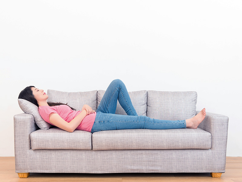 Mulher de camiseta rosa e calça jeans deitada em um sofá cinza no pós-operatório da mamoplastia de redução
