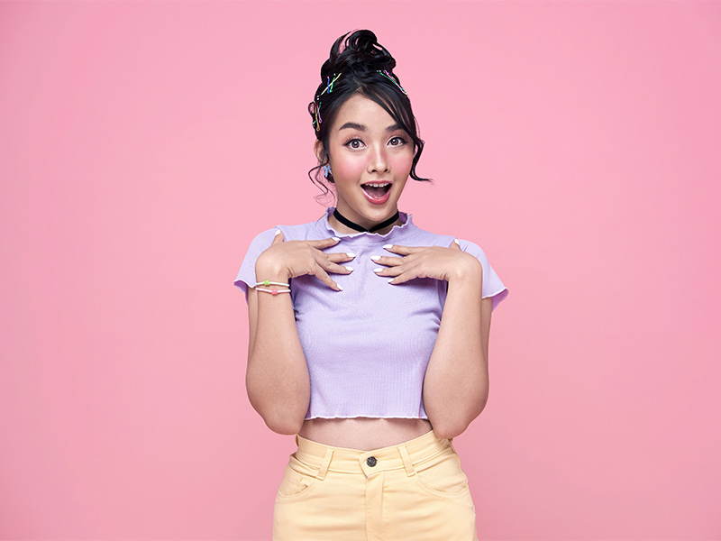 Com quantos anos pode fazer a mamoplastia redutora? Menina de cropped roxo, calça amarela e coque no cabelo, com cara de chocada com a idade que pode fazer a redução das mamas