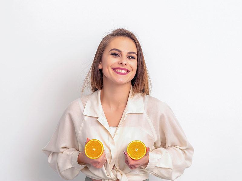 Mulher sorrindo de camisa branca, segurando duas laranjas cortadas na frente dos seios, para mostrar que a sensibilidade dos mamilos retorna após a mamoplastia