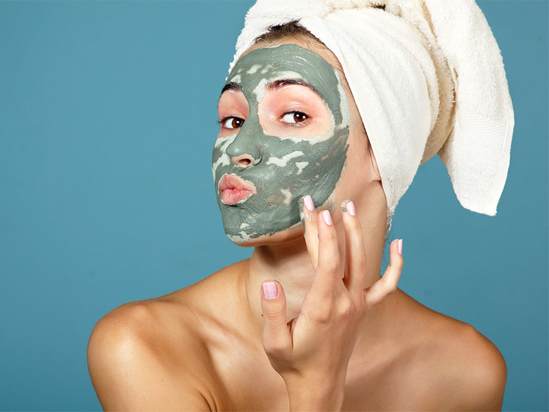 Imagem de uma mulher usando uma toalha branca na cabeça e uma máscara facial de argila verde