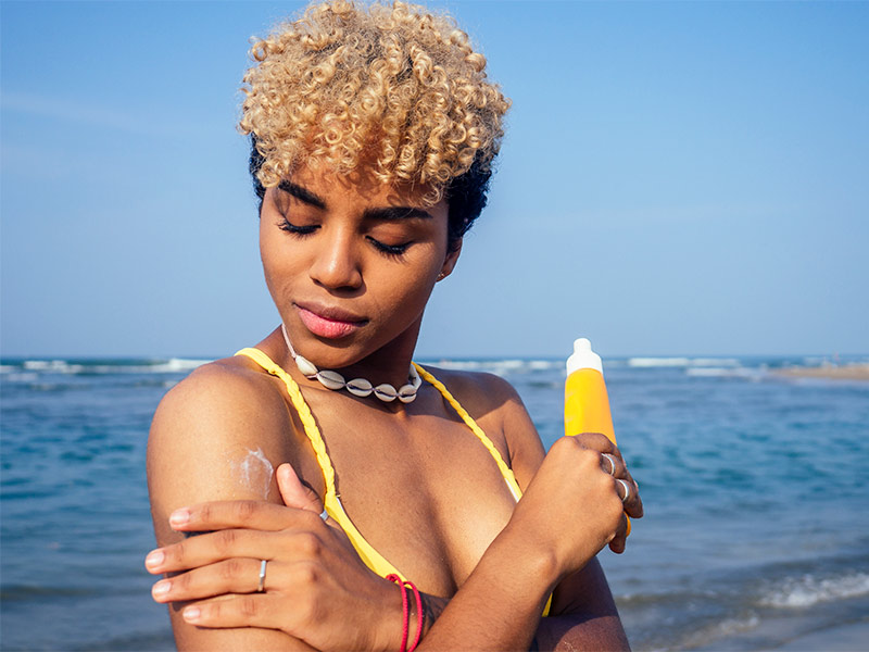 Quanto tempo depois da redução de mamas pode pegar sol? Mulher passando protetor solar no braço na praia para tomar sol depois da redução de mamas